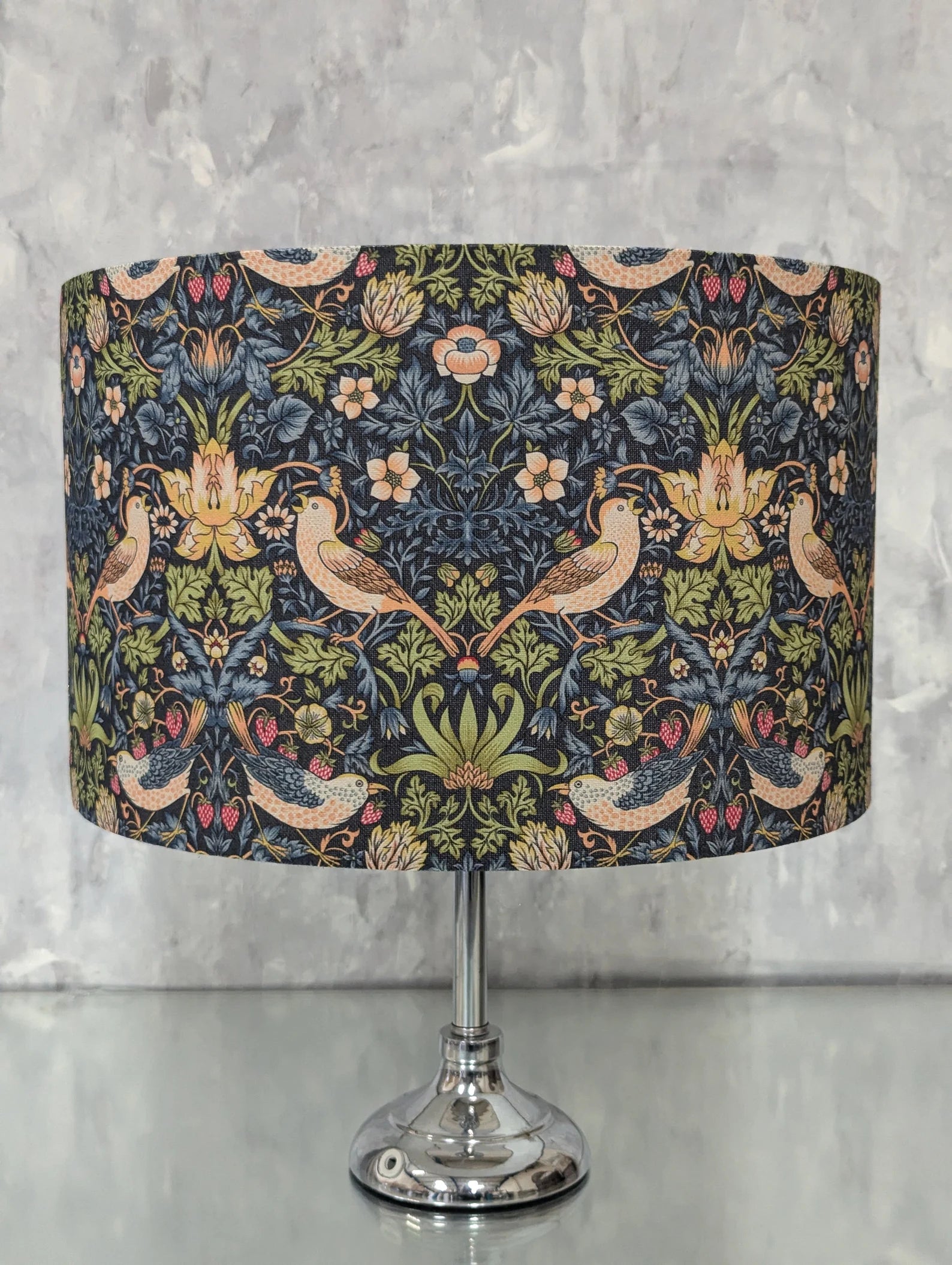 A table lamp with a dark navy blue fabric shade featuring a detailed William Morris-style floral and bird pattern in green, beige, and pink, mounted on a polished silver metal stem and base.