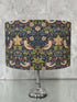A table lamp with a dark navy blue fabric shade featuring a detailed William Morris-style floral and bird pattern in green, beige, and pink, mounted on a polished silver metal stem and base.
