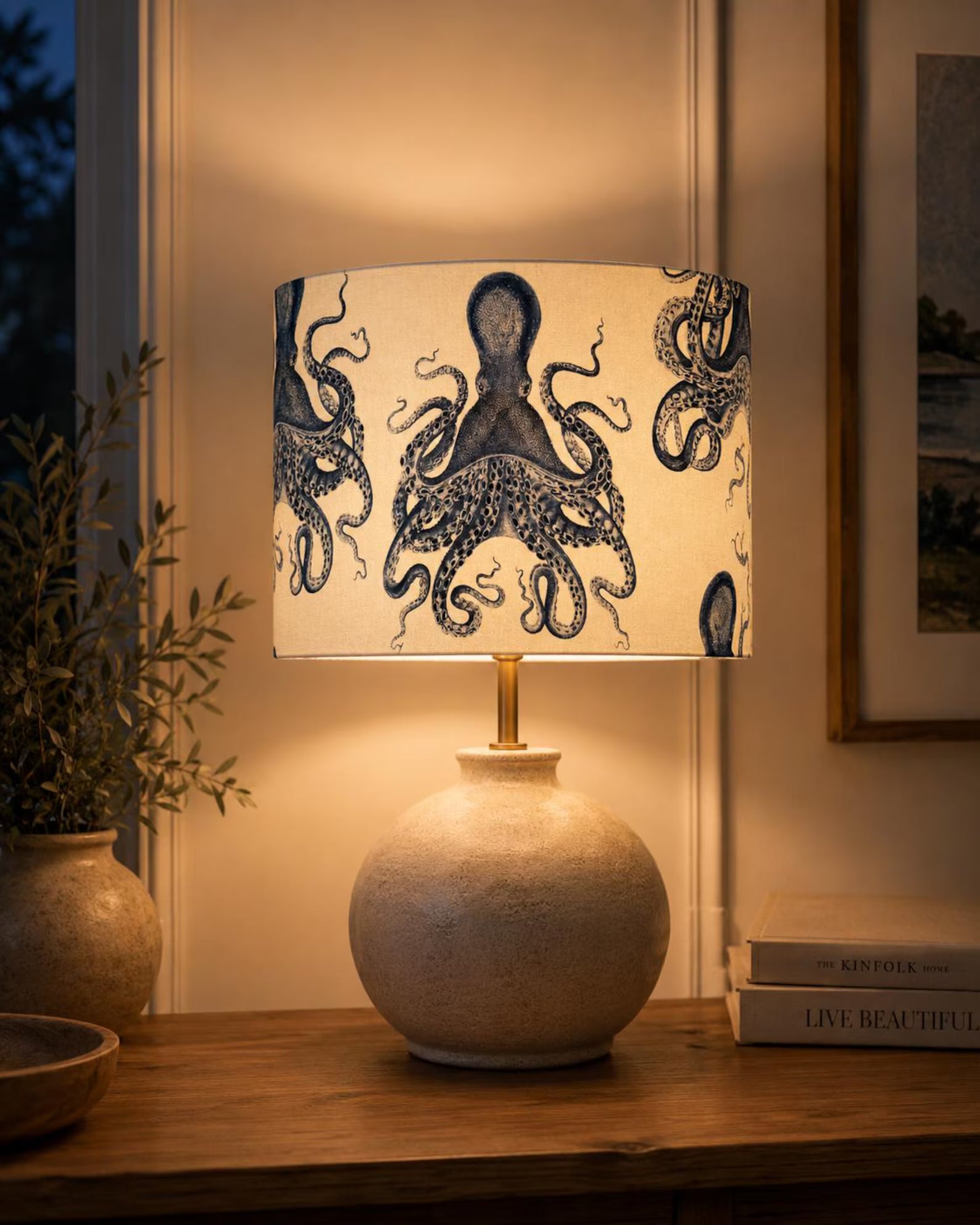 A table lamp with a textured cream ceramic base and a drum shade printed with navy blue octopus illustrations, lit warmly on a wooden table in a cozy interior setting.